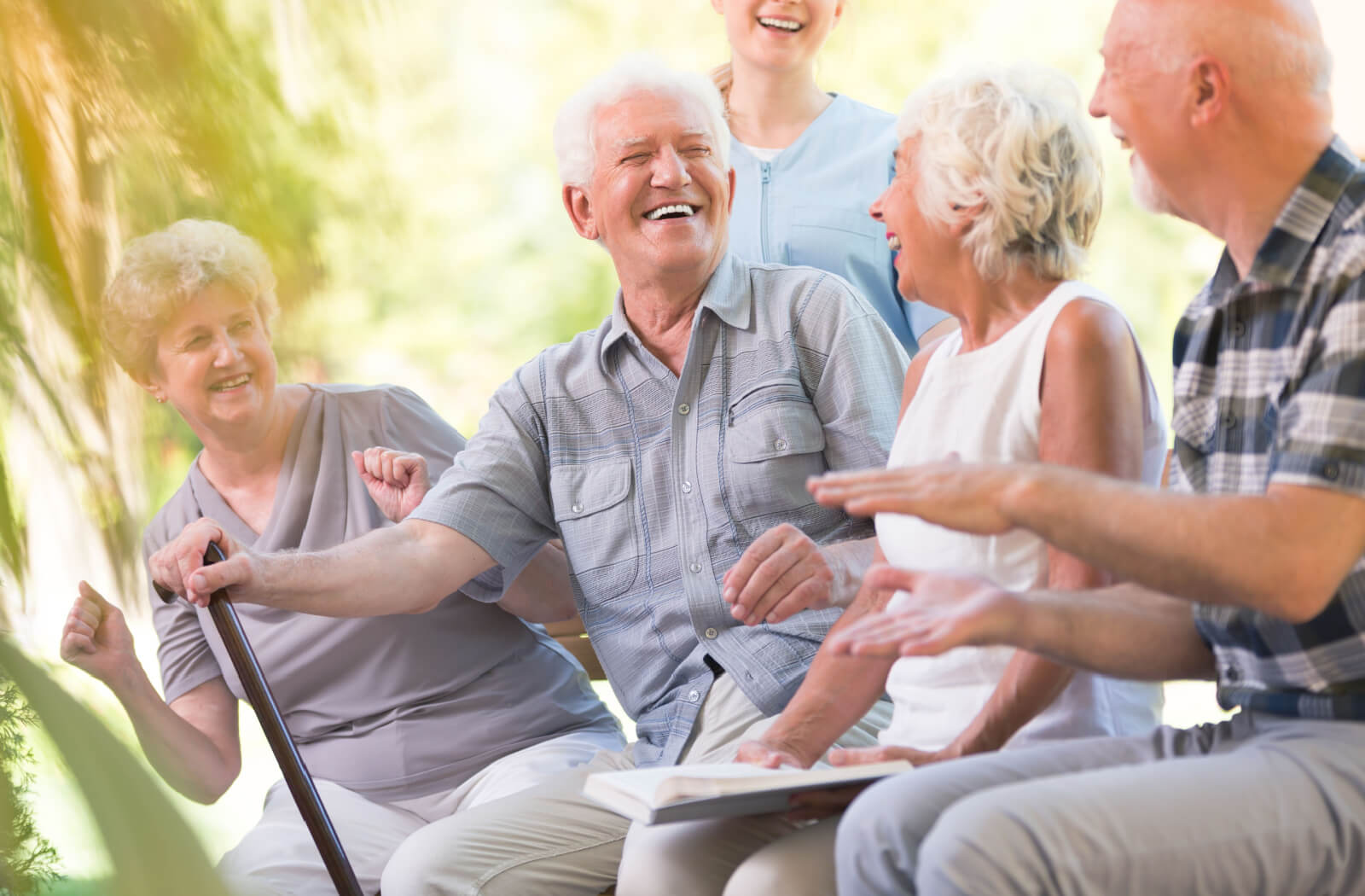 A senior man with a cane smiles and laughs with a nurse and other seniors.