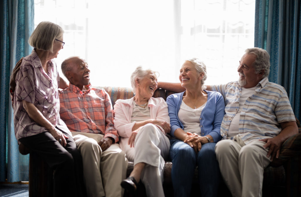 Group of smiling seniors talking, while sitting on couch against the window in senior living community