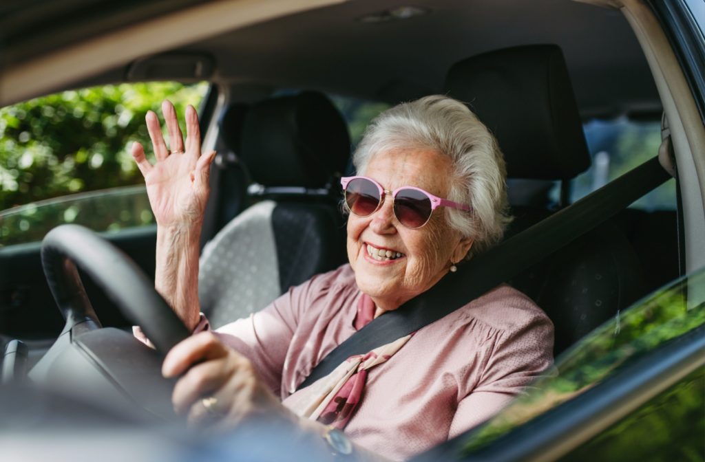 Elderly woman smiling and waving from the driver's seat.