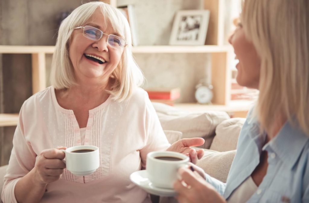 An older adult and their adult child sitting on a couch talking and laughing while they each hold a cup of coffee.