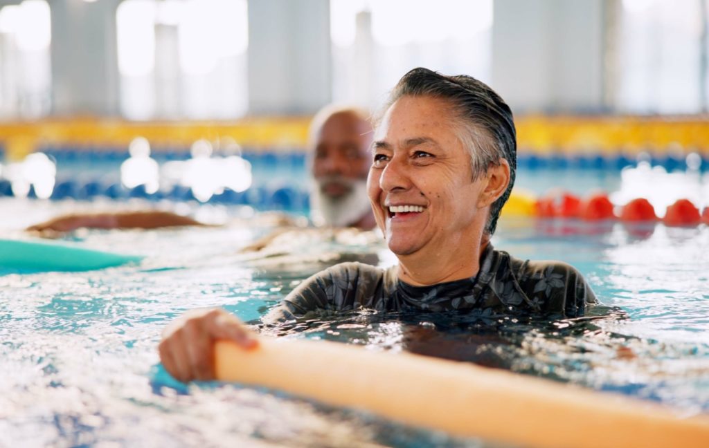 A senior in a water aerobics class smiles as they use an orange pool noodle to perform an exercise in the pool.