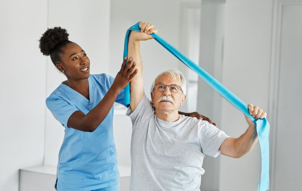 A senior does a strength training exercise with a blue resistance band while a nurse helps them with their form.