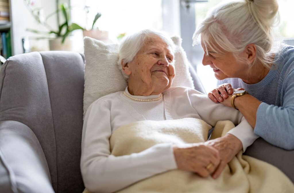 A smiling older adult lying down in respite care while their adult child holds their hands and smiles during a visit.